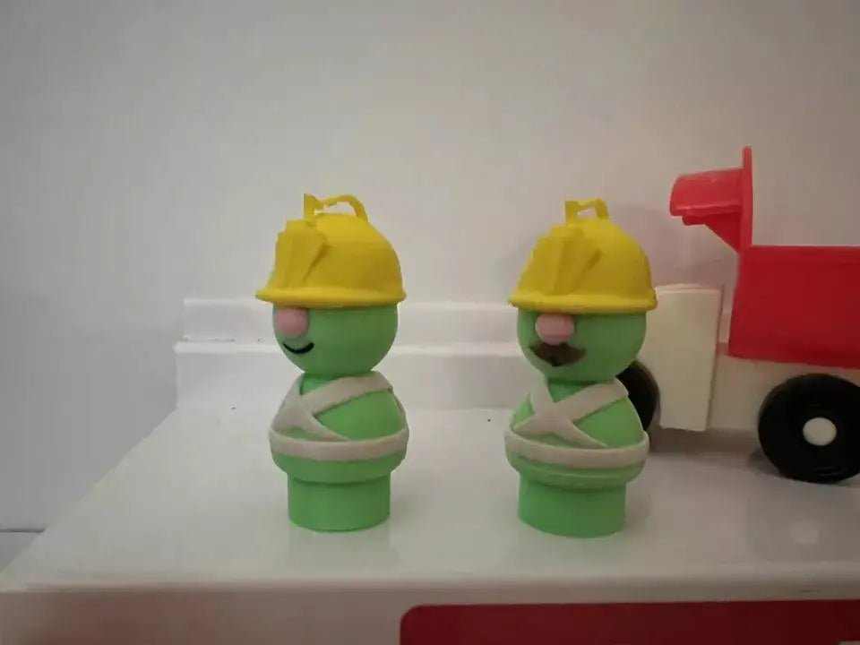 Two standing green Doozer toy figures with yellow hats beside a red cart