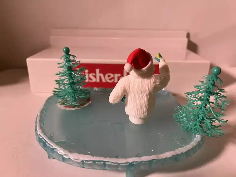 Holiday yeti decoration standing on gray base with red hat and green pine trees